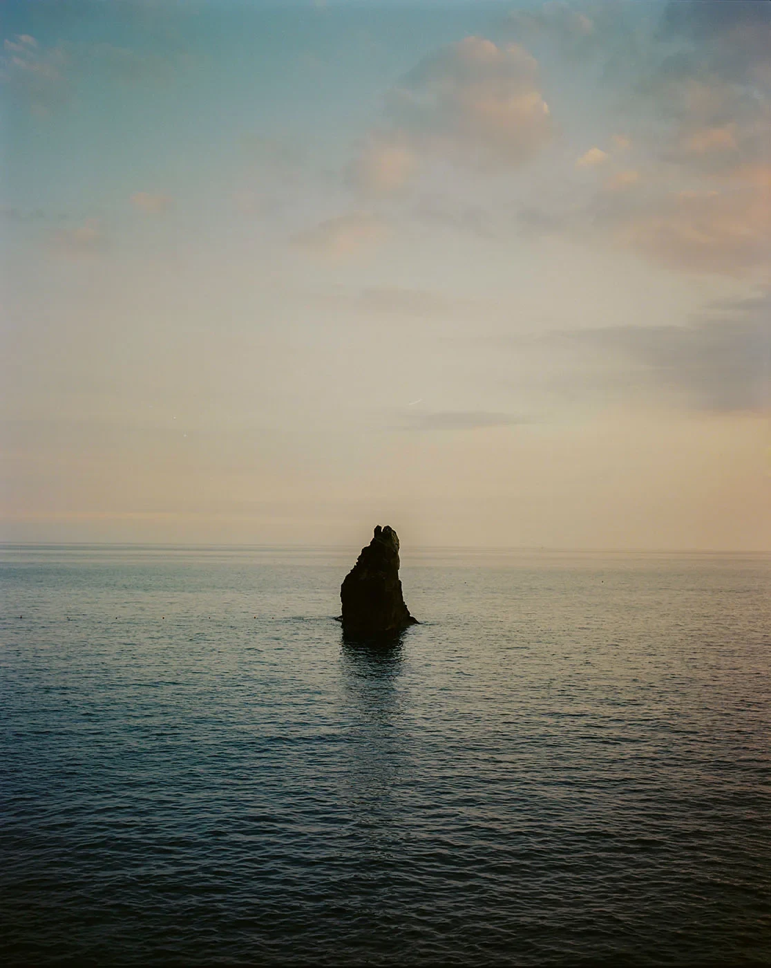 A solitary rock rises from a calm sea beneath a pale sky.