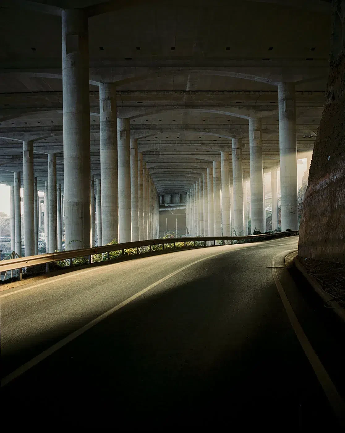 An empty curved road passes beneath repeated concrete columns, crossed by a band of sunlight.
