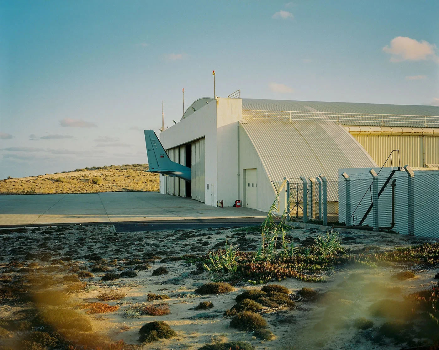 A pale industrial building stands beside wet ground and a metal fence.