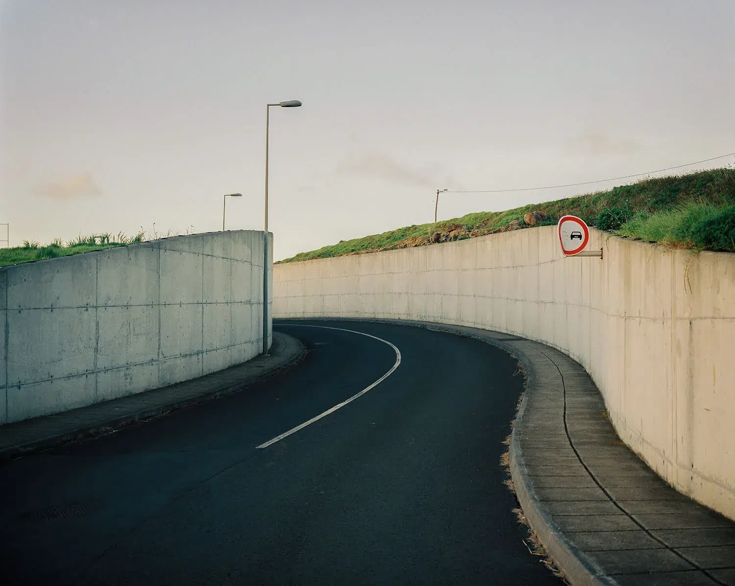 Uma estrada vazia curva entre muros altos de betão, sob candeeiros de rua.