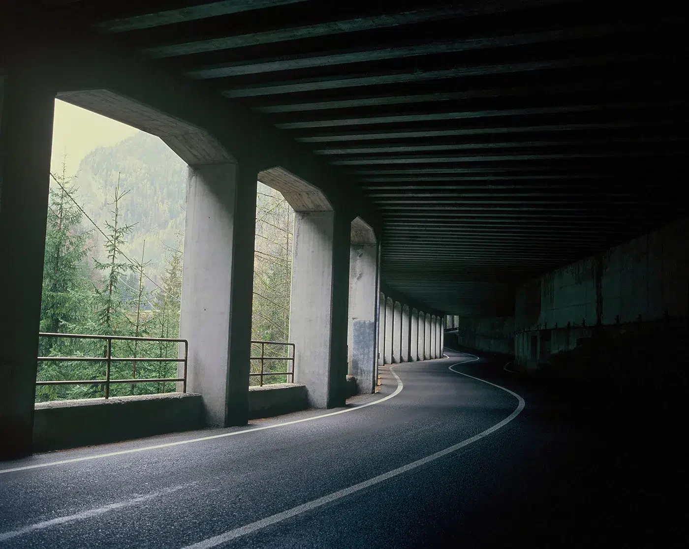 Uma estrada curva numa passagem coberta escura, com árvores visíveis entre as colunas.