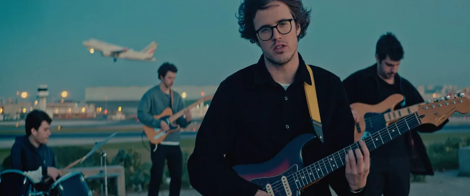 Uma banda a tocar no exterior, com um avião a passar ao fundo.
