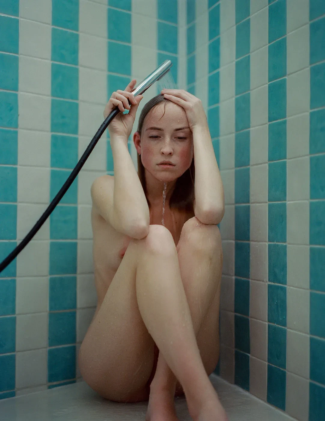 A person sits in a shower lined with turquoise tiles, holding a shower head above their head.