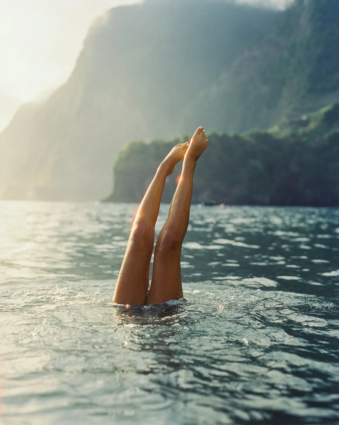 Two legs rise out of the water beside a mountainous shoreline.