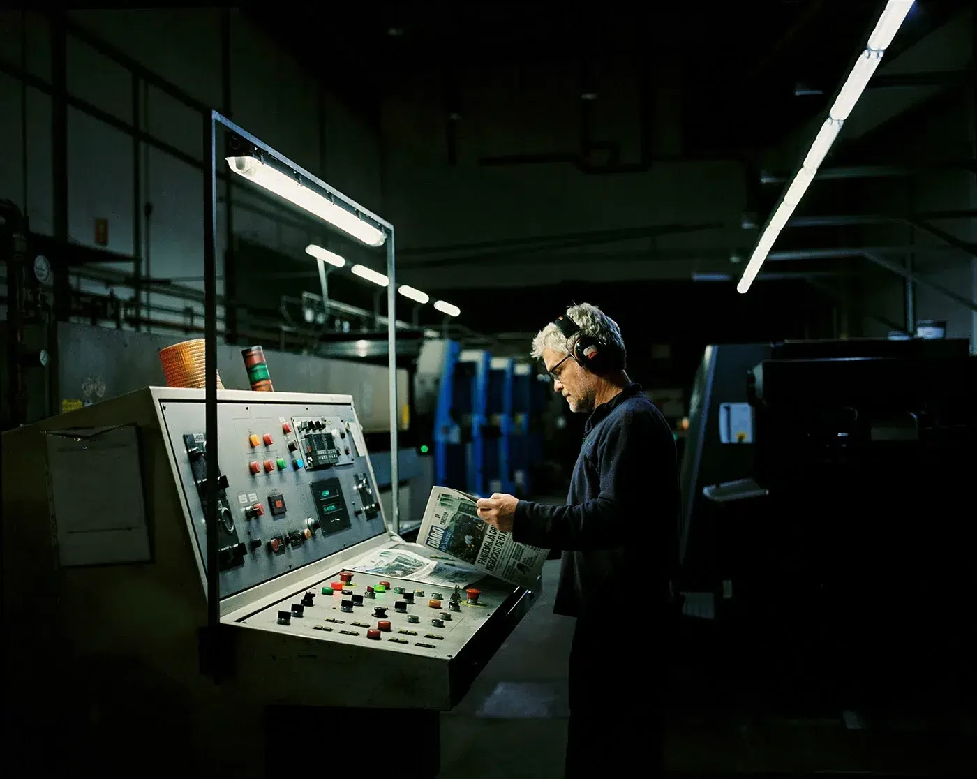 Uma pessoa está junto a um painel de controlo entre máquinas industriais, sob luzes fluorescentes.