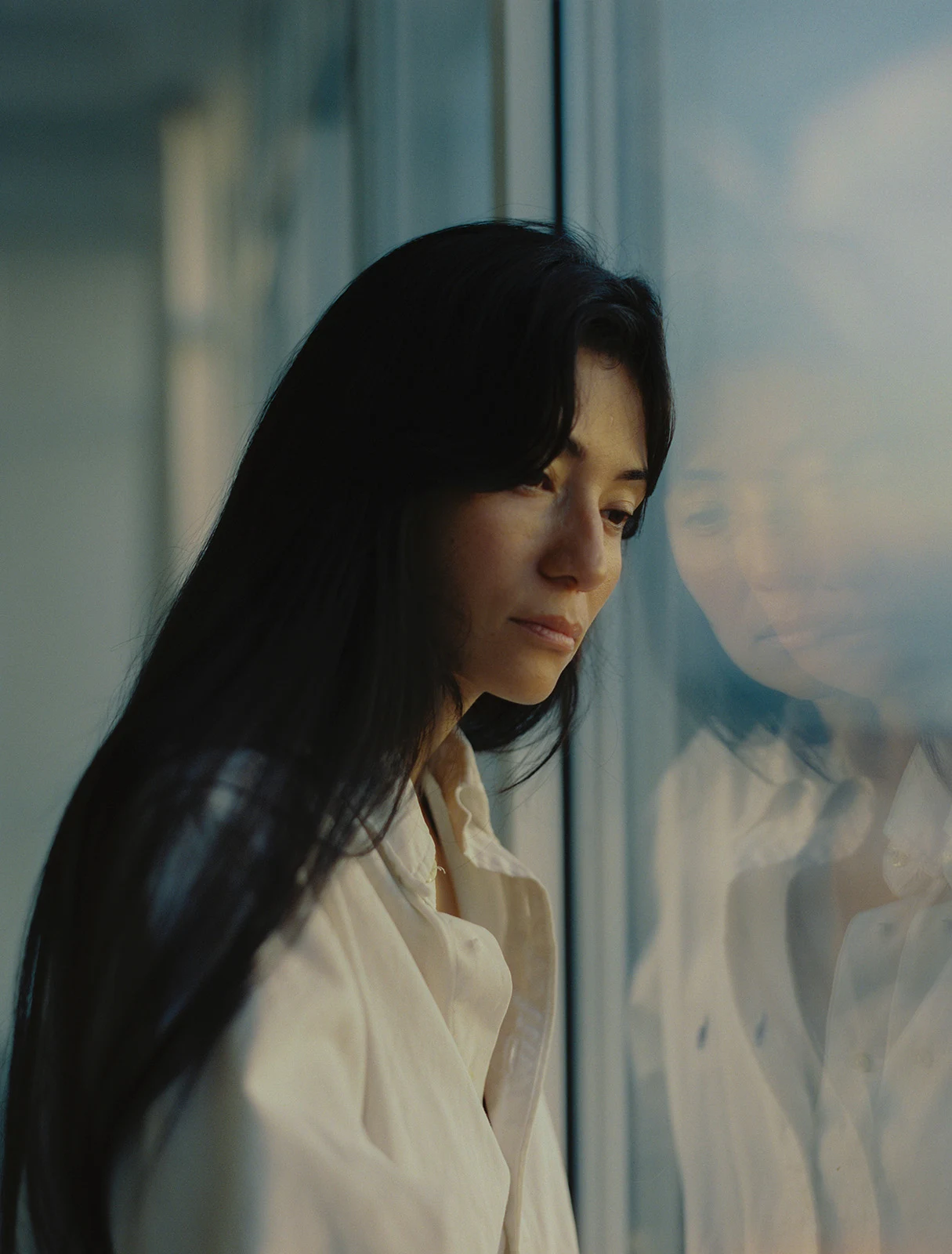 A person with long dark hair stands beside a window, with their reflection visible in the glass.