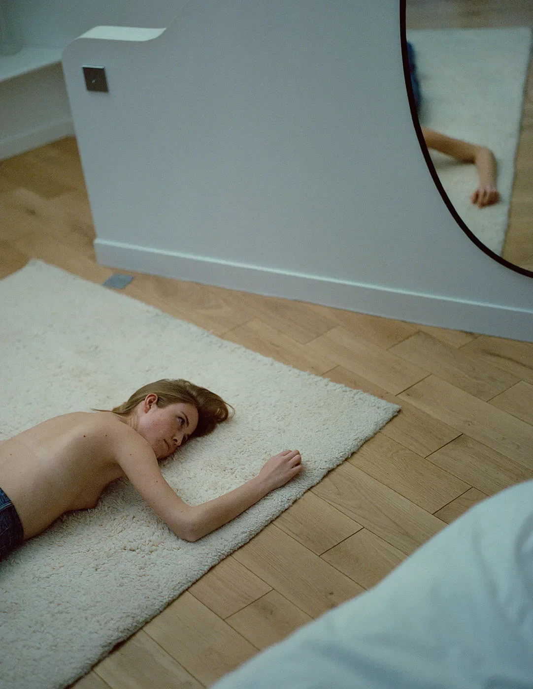 A person lies on a pale rug on a wooden floor beside a curved mirror.