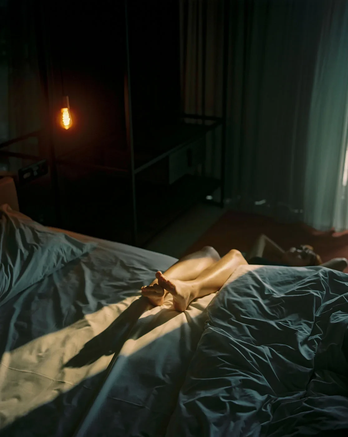 Sunlight falls across a person's legs on a bed in a dark room with a glowing hanging bulb.
