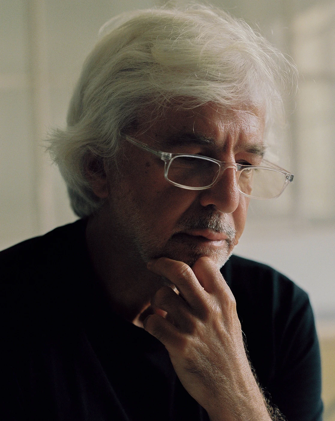A white-haired person wearing glasses rests their chin on one hand beside a window.