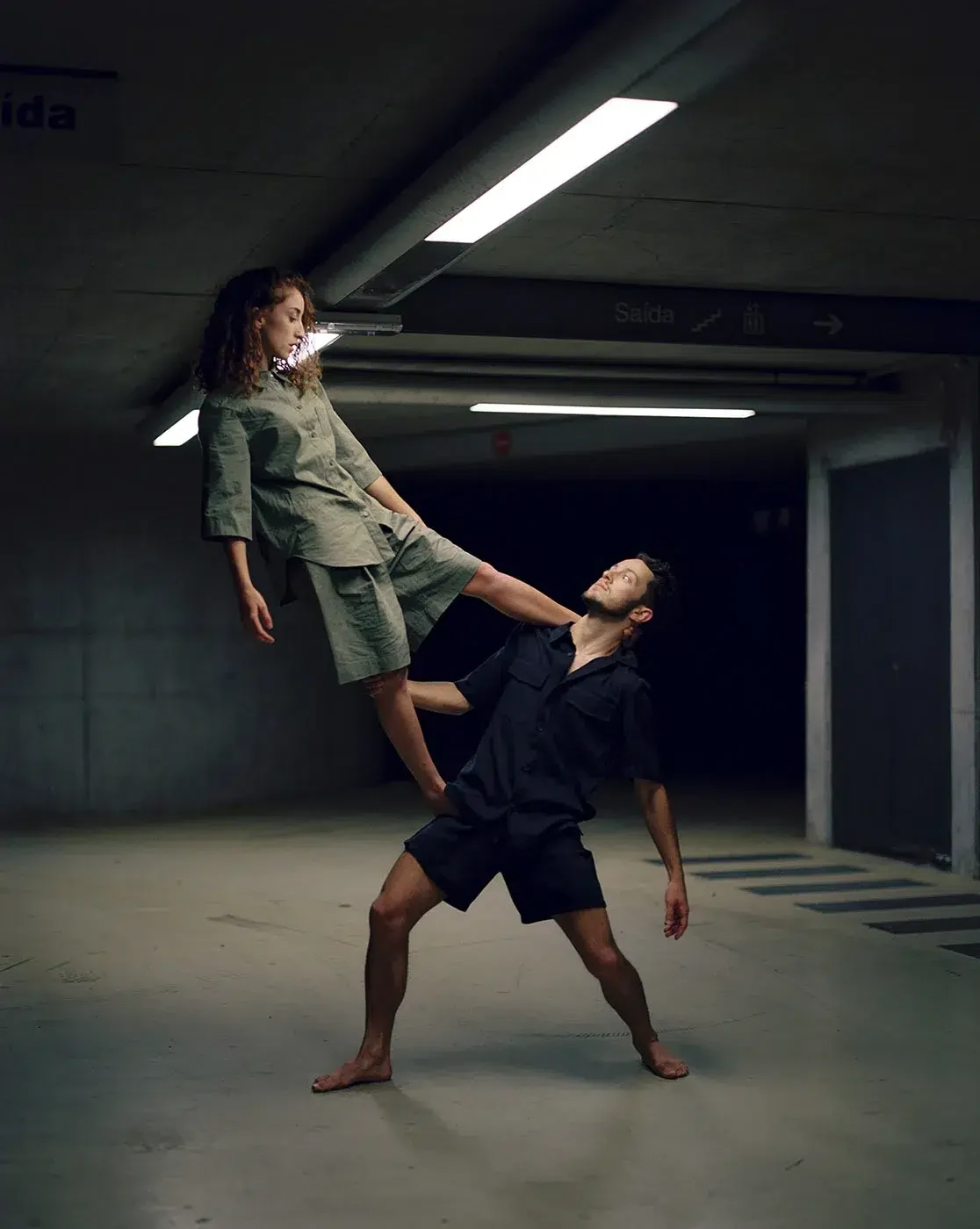 Duas pessoas descalças numa pose de dança com elevação, sob lâmpadas fluorescentes num interior de betão.