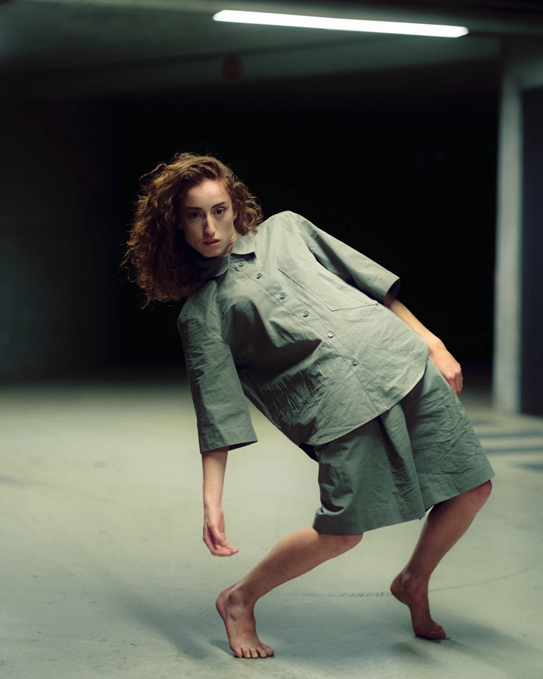 A barefoot person in a grey shirt and shorts leans sideways in a sparsely lit room.