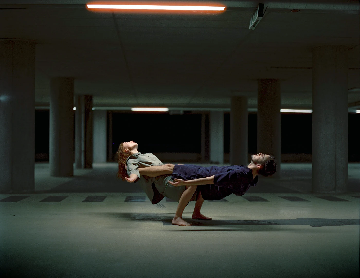 Duas pessoas descalças inclinam-se para trás em direções opostas numa pose de dança, num espaço de betão.