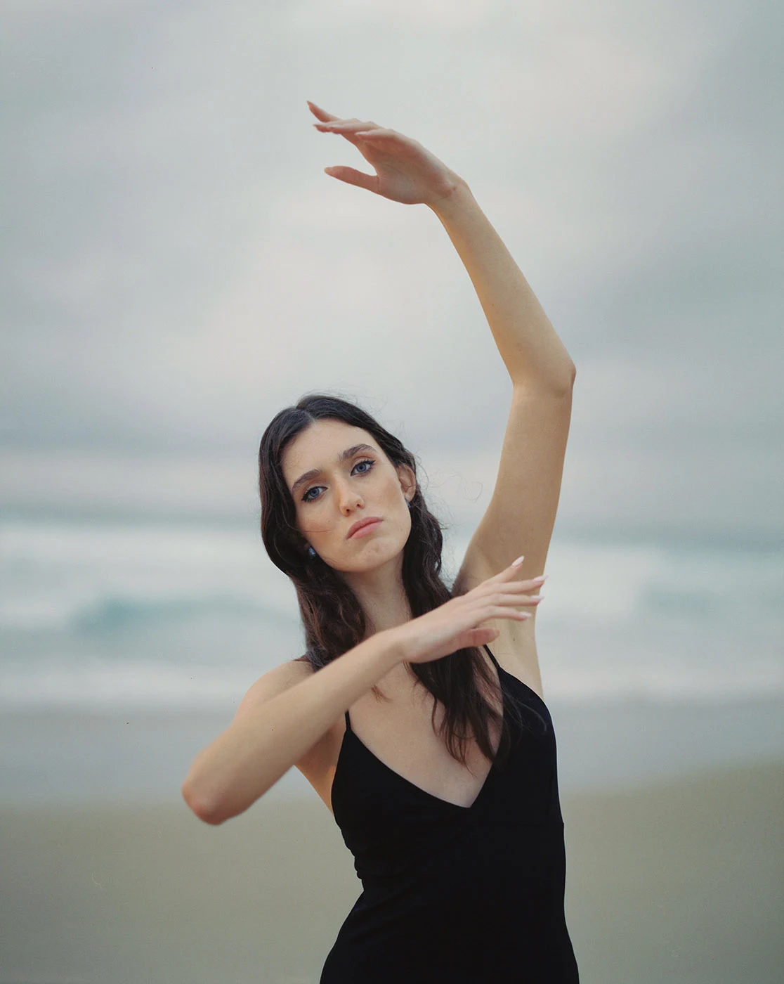 Uma pessoa vestida de preto ergue um braço numa pose de dança na praia.