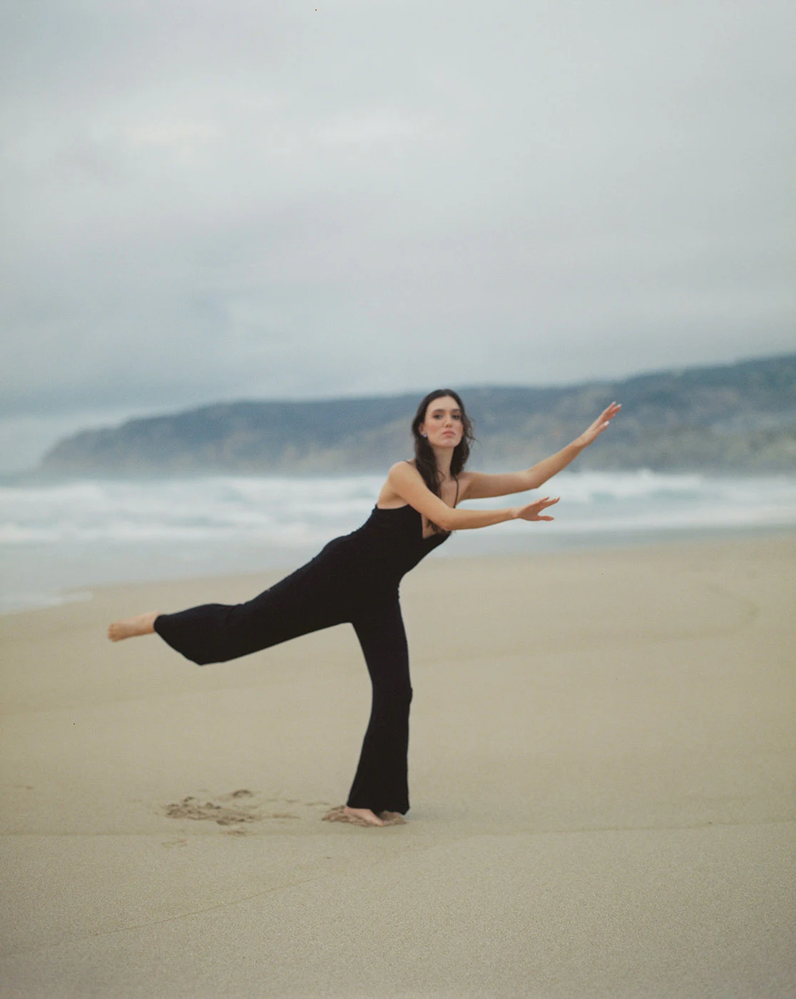 Uma pessoa equilibra-se numa perna numa pose de dança na areia, com ondas ao fundo.