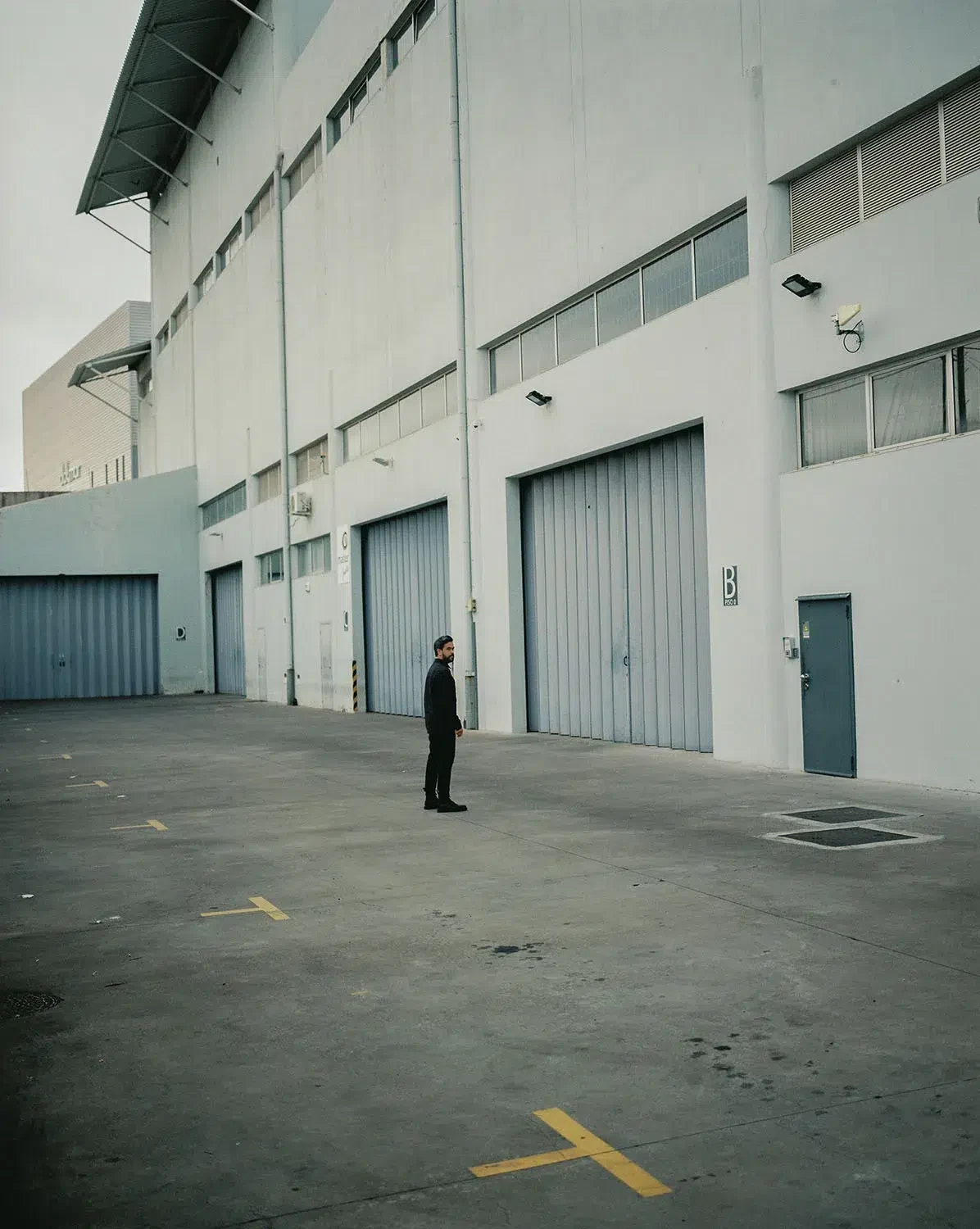 A lone person stands in a large paved yard beside a white industrial building.