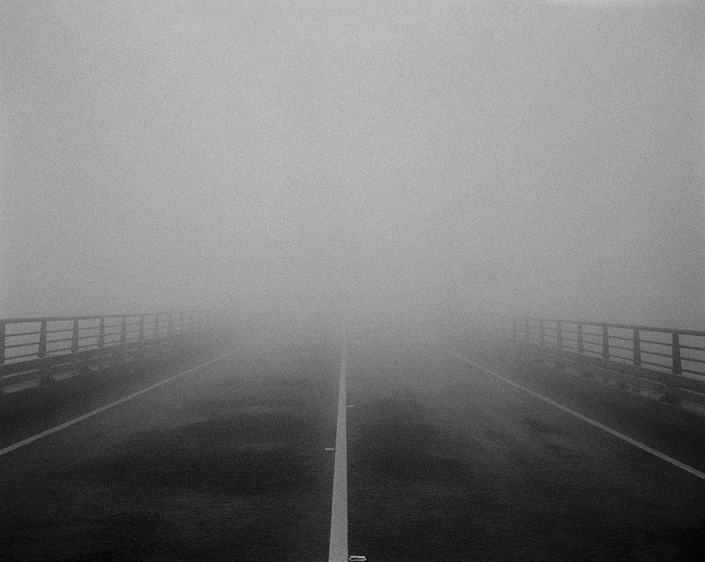 Vista a preto e branco de uma estrada vazia que desaparece no nevoeiro denso entre corrimãos.