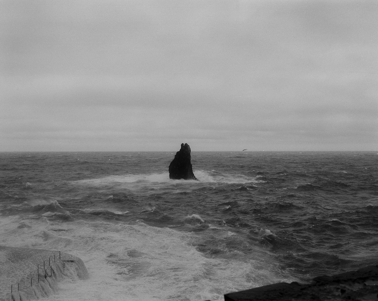 Vista a preto e branco de um rochedo isolado no mar, rodeado por ondas.
