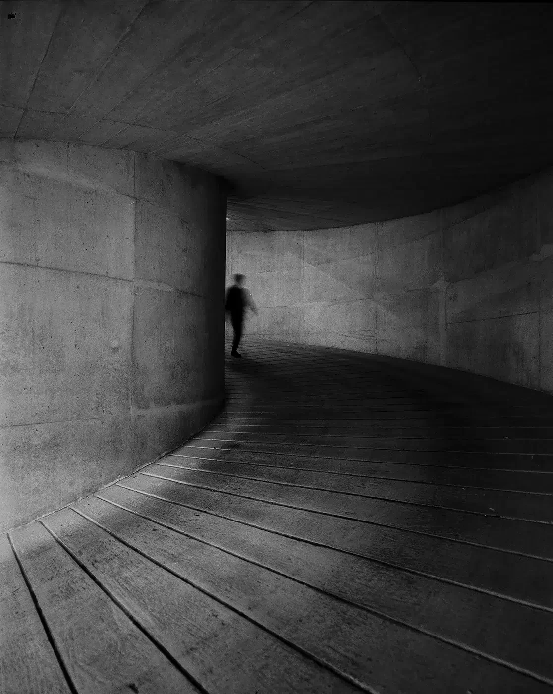 Fotografia a preto e branco de uma figura distante numa passagem curva de betão com pavimento de madeira.