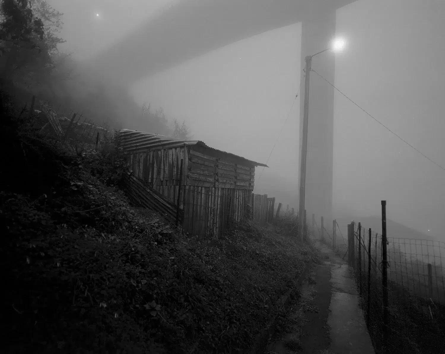 Vista a preto e branco de um caminho estreito e uma pequena construção sob um viaduto envolto em nevoeiro.